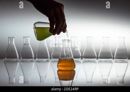 Hand Gießen grün in orange Flüssigkeit in wissenschaftlichen Labor Glas erlenmeyerkolben mit Glasgeräten auf reflektierende Oberfläche. Stockfoto