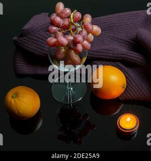 Komposition aus Trauben in einer Glasschale, Orangen, einem Wollschal und einer brennenden Kerze auf einem reflektierenden, dunkel glänzenden Hintergrund Stockfoto