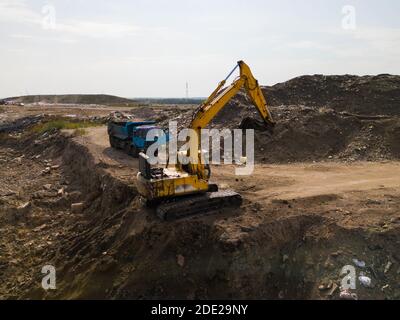 Schwere Ausrüstung auf Aushubgrube. Gelber Bagger und blauer LKW auf unbefestigter Straße. Haufen von Sand und Steinen. Stockfoto