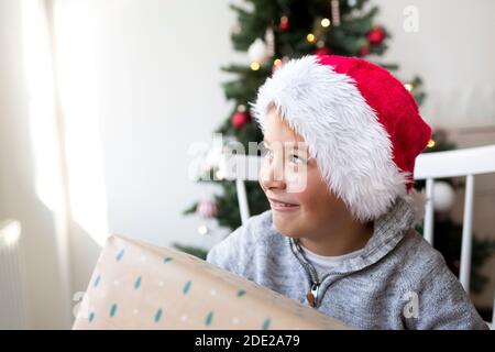 Kleiner Junge in santa Klausel Hut vor einem Weihnachtsbaum mit Weihnachtsgeschenken Stockfoto