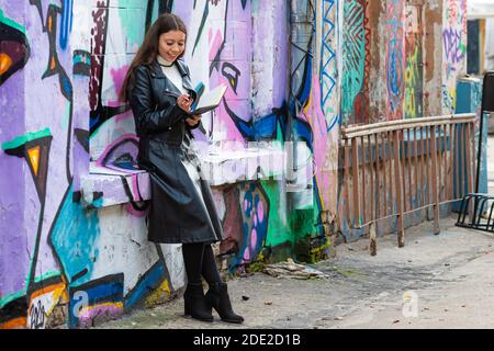 Riga, Lettland - 17. Oktober 2020: Eine junge Frau lehnt sich an eine mit Graffiti besprühte Wand und macht Notizen in einem schwarzen Notizbuch Stockfoto