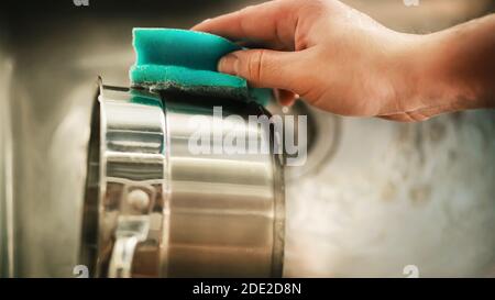 Ein Mann hält einen seifigen blauen Schwamm in der Hand und wäscht ihn, um eine Metallpfanne über dem Spülbecken zu waschen. Hausarbeit. Küchenutensilien. Stockfoto