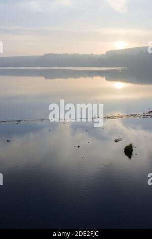 Neblige Landschaft und wispige Wolken spiegeln sich in ruhigem Reservoirwasser. Vertikales Sonnenuntergangsbild über dem Rotmires-Reservoir am oberen Reservoir. Stockfoto