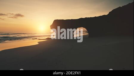 Sonne Silhouette der Felswand mit riesigen Loch auf Sand Strand Luftbild. Sandige Ozeanküste, einzigartige Klippe auf Sumba Island, Indonesien, Asien. Niemand Natur bei Sonnenlicht. Filmischer Sonnenuntergang im Sommer Stockfoto