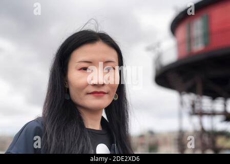Eine chinesische Frau, die in der Nähe des 7 Fuß hohen Leuchtturm lächelt, verschwimmt im Hintergrund mit einem grau bewölkten Tag in Baltimore Maryland. Stockfoto