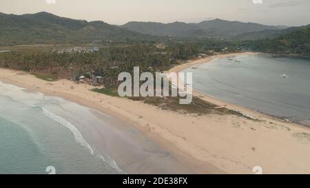 Tropisches Resort mit Touristen am Sandstrand Luftaufnahme. Ozeanküste mit Resort, beherbergt Gebäude an grünen Palmen. Reisende am Meer. Schiffe, Schiffe, Boot in seichtem Wasser im Hafen von Philippinen Stockfoto