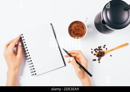 Draufsicht weibliche Hände schreiben in leerem Papier Notebook neben Kaffeetasse und Geysir-Kaffeemaschine auf weißem Tisch. Stockfoto