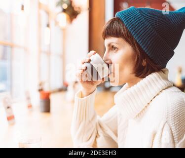 Reife Frau trinkt Kaffee und schaut aus dem Fenster, kaltes Wetter, warme Kleidung Stockfoto