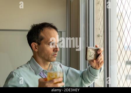 44-jähriger Mann, vor einem Fenster, nimmt einen Whisky und schaut sich Videos auf seinem Smartphone an. Stockfoto