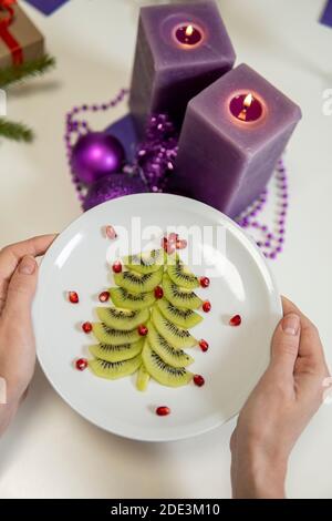 weihnachtsdekoration. Kiwi und Granatapfel in Form eines weihnachtsbaums auf einem Teller und lila Kerzen. Hohe Qualität Stockfoto