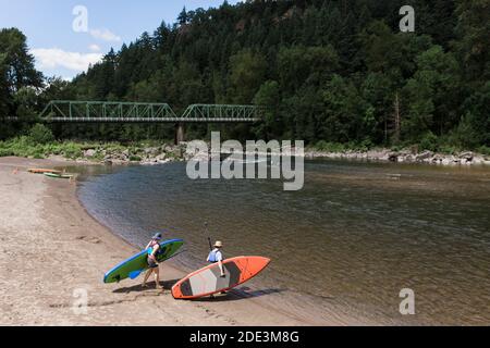 Ein junges Paar trägt Standup-Paddleboards zum Wasser in Oregon. Stockfoto