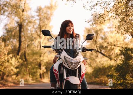 Lächelnde selbstbewusste junge Frau auf dem Motorrad auf Landstraße sitzen Inmitten von Bäumen Stockfoto