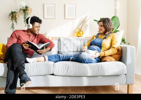 Schwangere Frau, die mit ihrem Mann, der Musik hört und ein Buch liest, auf der Couch isst. Interracial paar Konzept Stockfoto