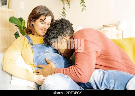 Ehemann küsst seine schwangere Frau auf einem Sofa. Interracial paar Konzept Stockfoto