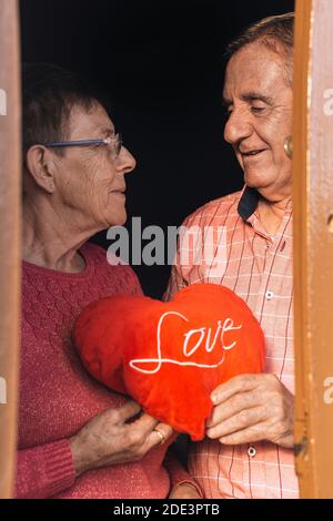 Lächelndes, romantisches Senioren-Paar, das rotes Herz hält und anschaut Einander mit Liebe, während die Darstellung glückliche Ehe und Beziehung Stockfoto