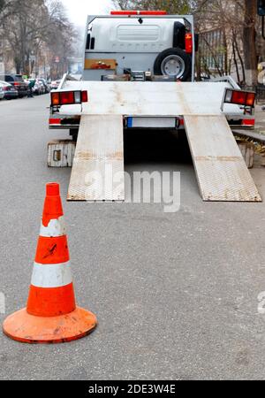 Ein leuchtend orangefarbener Verkehrskegel steht auf dem dunklen Asphalt und zäunt die Fahrbahn mit einem geparkten Transportwagen, vertikales Bild, Kopierraum. Stockfoto