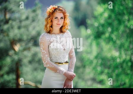 Schöne junge rothaarige Braut im Wald mit einem Blumenkranz auf dem Kopf. Frau in langen weißen Kleid im Freien am Sommertag. Hochzeitstag. Stockfoto