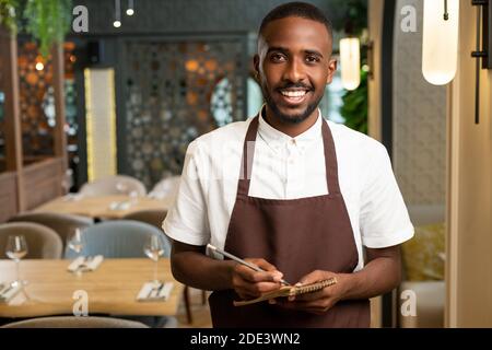 Glücklicher junger Kellner mit toothy Lächeln halten Stift und klein Notepad beim Stehen vor der Kamera im Restaurant und Bestellungen werden entgegengenommen Stockfoto