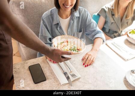 Junge lächelnde Frau in blauem Hemd sitzen am Tisch während Afrikanischer Kellner bringt Teller mit Salat und legt ihn auf Tisch, während sie serviert Stockfoto