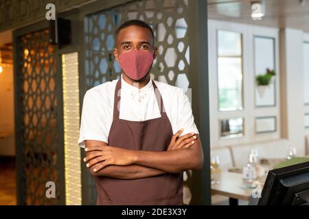 Moderner Kellner in Schutzmaske und brauner Schürze, die Arme kreuzt Auf der Brust beim Stand am Computer in modernen Café oder Restaurant Stockfoto