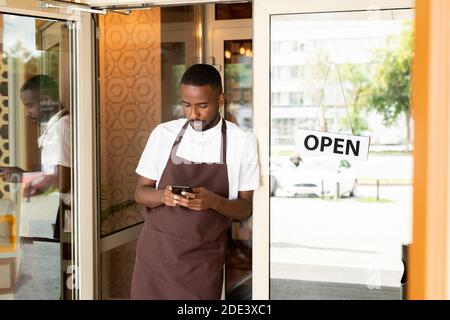 Afrikanischer junger Kellner in Uniform steht in der Tür von Café oder Restaurant und Scrollen durch neue Aufträge von Kunden Auf dem Smartphone Stockfoto