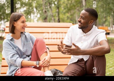 Glückliches junges interkulturelles Paar in Casualwear sitzen auf Holzbank im Park oder Platz, plaudern und genießen Sommertag in der Freizeit Stockfoto