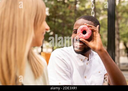Junge blonde Frau schaut und lacht auf lustig und fröhlich Afrikanischer Mann hält gespritzter Donut mit rosa Glasur von seinem Linkes Auge im Park Stockfoto