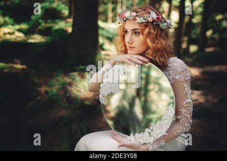 Schöne junge rothaarige Braut im Wald mit einem Blumenkranz auf dem Kopf und Spiegel. Frau in langen weißen Kleid im Freien am Sommertag. Hochzeit Stockfoto