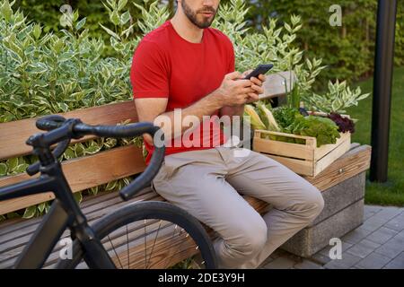 Männlicher Kurier in Uniform sitzt auf der Bank im Freien und mit mobilen App während der Lieferung Lebensmittelkarton mit frischem Gemüse auf dem Fahrrad. Konzept für die Lebensmittelzustellung Stockfoto