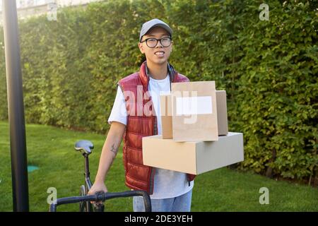 Junge asiatische männliche Kurier hält Pakete und Papiertaschen und Boxen für die Lieferung und Blick auf Kamera, während mit seinem Fahrrad im Freien stehen. Konzept der Delivery Services Stockfoto