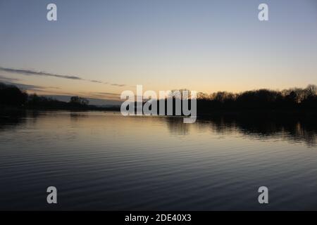 Blick bei Sonnenuntergang mit blauem Himmel mit einigen Wolken am Horizont, die sich an einem kalten Herbsttag auf einem Seewasser spiegeln, intensive orange und dunkle Farben in der Dämmerung Stockfoto