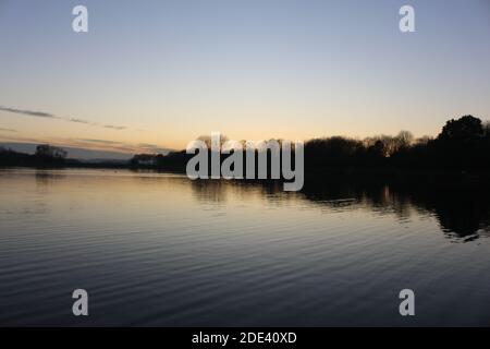 Blick bei Sonnenuntergang mit blauem Himmel mit einigen Wolken am Horizont, die sich an einem kalten Herbsttag auf einem Seewasser spiegeln, intensive orange und dunkle Farben in der Dämmerung Stockfoto