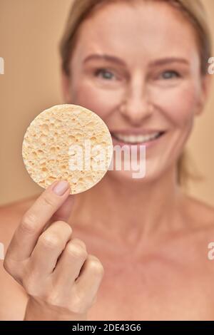 Vertikale Aufnahme einer charmanten reifen Frau halten Reinigung Make-up Schwamm im Stehen isoliert auf beigem Hintergrund im Studio Stockfoto