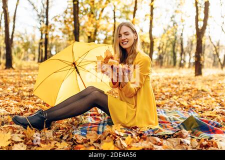 Ein junges, glückliches Mädchen in einem Herbstpark sitzt auf einer Decke, hält Herbstblätter und genießt das warme sonnige Wetter. Das Konzept der Herbstsaison Stockfoto