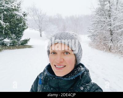 Glücklich lächelnde kaukasische Frau, die Selfie-Foto auf dem Smartphone im Park im Freien am Wintertag. Starker Schneefall und Schneesturm. Erster Frostschnee. Schönheit Stockfoto