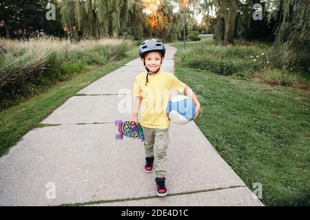 Happy Kaukasischen Jungen im Helm hält Ball und Skateboard im Park am Sommertag. Saisonale Outdoor-Aktivitäten für Kinder Sport. Gesunder Lebensstil im Kindesalter Stockfoto