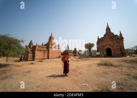 Buddhistischer junger Mönch in roter Robe steht mit Schirm vor einem Tempel, Bagan, Myanmar Stockfoto