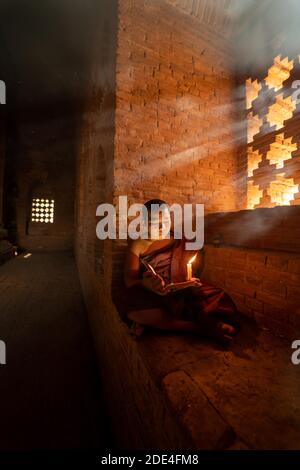 Buddhistischer junger Mönch in roter Robe, der vor Lichtstrahlen in einem Tempel in Bagan, Myanmar, sitzt Stockfoto