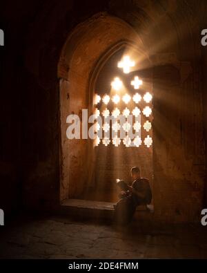 Buddhistischer junger Mönch in roter Robe, der vor Lichtstrahlen in einem Tempel in Bagan, Myanmar, sitzt Stockfoto