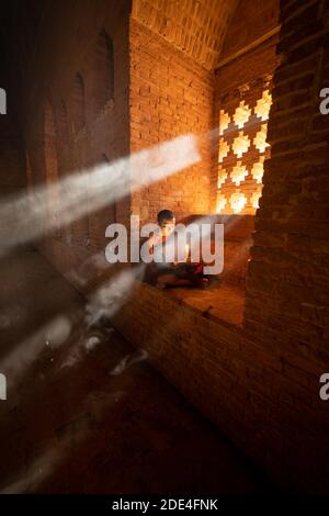 Buddhistischer junger Mönch in roter Robe, der vor Lichtstrahlen in einem Tempel in Bagan, Myanmar, sitzt Stockfoto