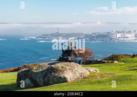 Blick von hinten auf Dudelsackläufer, der auf einem Felsen sitzt und spielt Die Dudelsäcke mit dem Turm von Hecules im Hintergrund Im Nebel Stockfoto