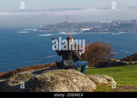 Blick von hinten auf Dudelsackläufer, der auf einem Felsen sitzt und spielt Die Dudelsäcke mit dem Turm von Hecules im Hintergrund Im Nebel Stockfoto