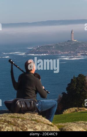 Blick von hinten auf Dudelsackläufer, der auf einem Felsen sitzt und spielt Die Dudelsäcke mit dem Turm von Hecules im Hintergrund Im Nebel Stockfoto