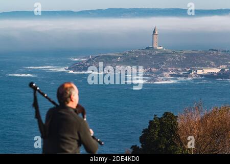 Blick von hinten auf Dudelsackläufer, der auf einem Felsen sitzt und spielt Die Dudelsäcke mit dem Turm von Hecules im Hintergrund Im Nebel Stockfoto