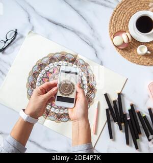 Frau macht Fotos von farbigen Mandala aus Skizzenbuch auf Schreibtisch aus weißem Marmor mit ausgestellten Objekten Stockfoto