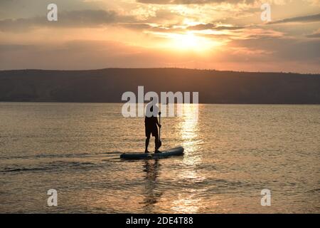 Mann segelt auf einem SUP-Board in einem großen See bei Sonnenaufgang. Stand Up Paddle Boarding - aktive Erholung in der Natur. Der See von Galiläa, See Tiberias, Kinneret, Kinnereth. Hochwertige Fotos. Stockfoto