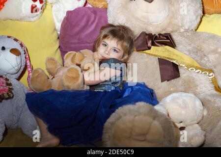 Ein kleines Mädchen in einem blauen Kleid liegt in einem Haufen von großen Plüsch Spielzeug Stockfoto