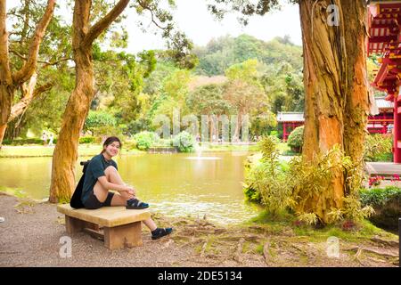 Biracial teen Mädchen ruht o Beton Bank neben ruhig Teich im asiatischen Stil in einem asiatischen Tempelgarten Stockfoto