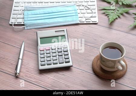 Holztisch mit Tastatur, Gesichtsmaske, schwarzem Kaffee, Stift, Rechner und grünen Pflanzen. Stockfoto