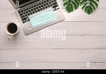 Schreibtisch aus Holz mit Laptop, Gesichtsmaske, Grünpflanzen und Kaffee. Draufsicht, Kopierbereich. Stockfoto